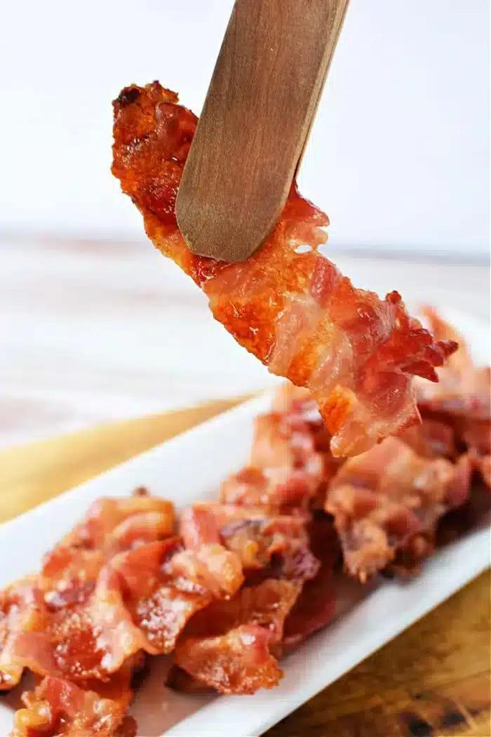 A piece of crispy bacon held by wooden tongs above a white rectangular plate filled with more bacon strips. The background is blurred, and the focus is on the bacon being lifted, showcasing perfectly cooked strips—just like what you'd get if you knew how to cook bacon in the oven with aluminum foil