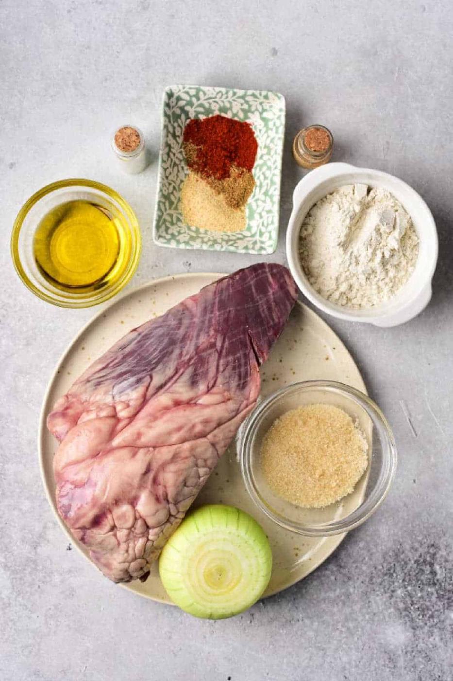 How to Cook Beef Heart: Raw beef heart, onion, flour, oil, brown sugar, and spices arranged on a light gray countertop.