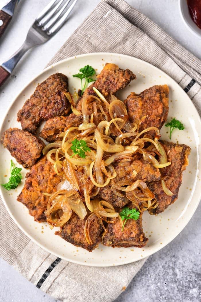 A plate of beef heart slices and onions garnished with fresh parsley features browned liver pieces alongside tender beef heart slices, all topped with sautéed onions. The dish rests on a cloth napkin with a fork and knife beside it.