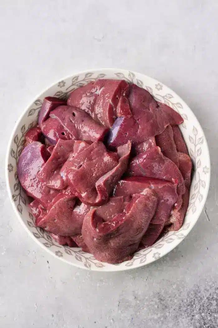 A white bowl filled with raw, sliced chicken liver sits on a light gray countertop.