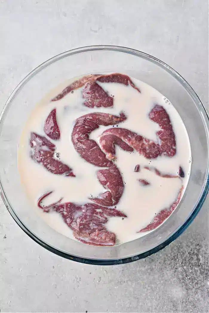 A glass bowl filled with pieces of raw liver soaking in milk rests on a light-colored, textured surface.