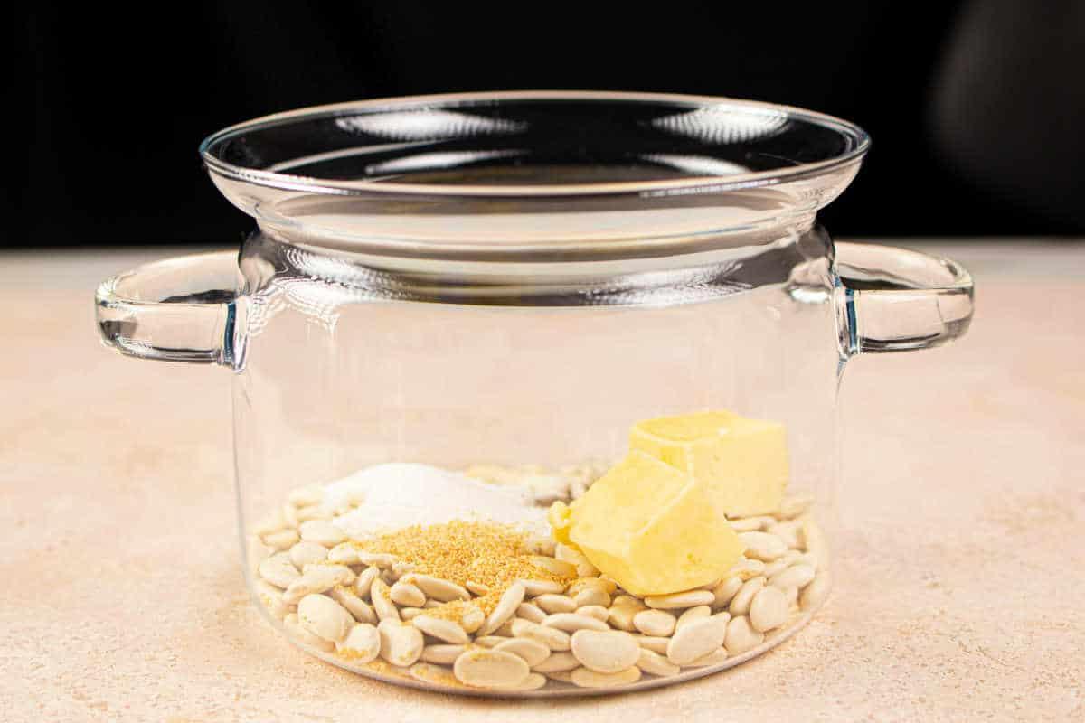 A glass pot with butter beans, butter, sugar, and spices inside rests on a light surface. Its reminiscent of preparing a comforting dish like butter beans—simple yet satisfying in its blend of ingredients.