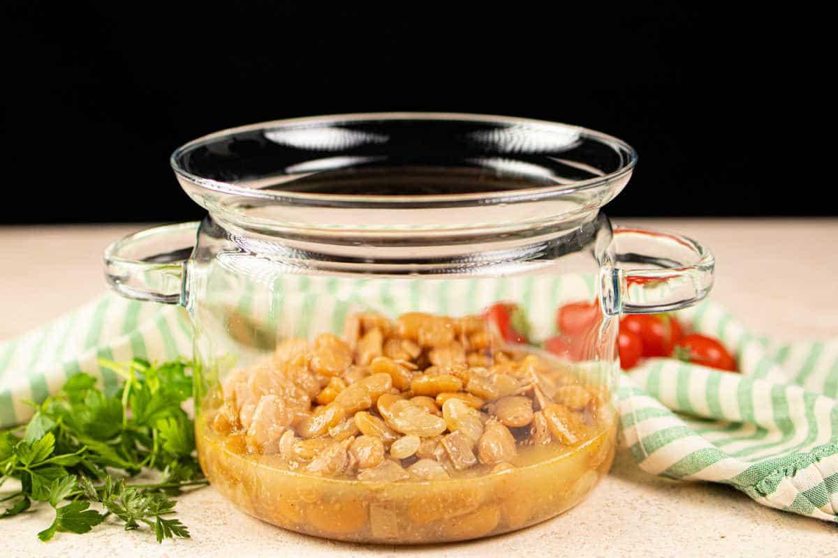 A glass pot filled with butter beans sits beside fresh parsley and cherry tomatoes on a striped cloth, offering inspiration on how to cook butter beans with a touch of garden-fresh simplicity.