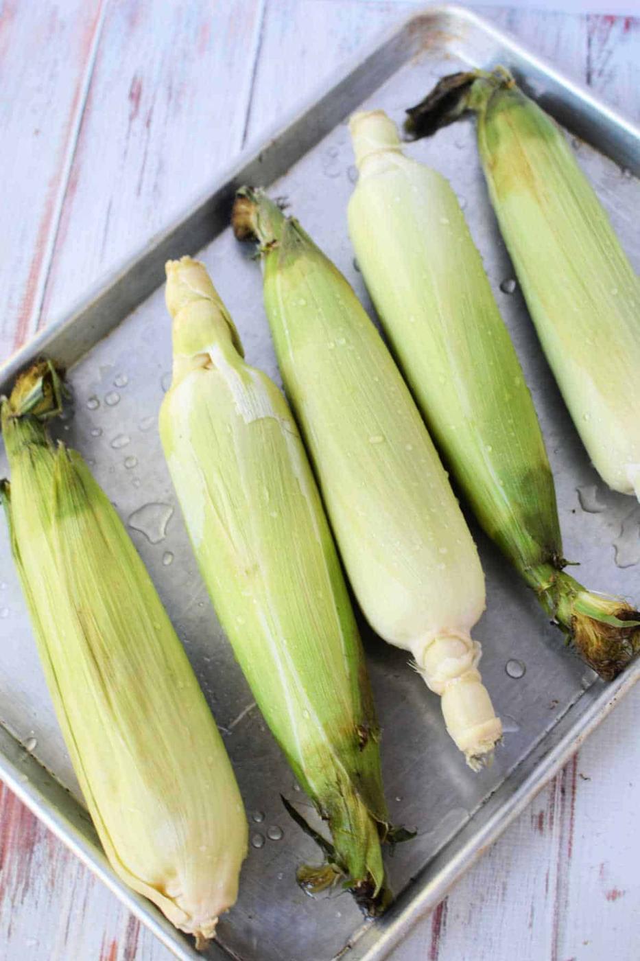 Five ears of corn in husks rest on a metal baking sheet, some glistening with water droplets—perfect for learning how to cook corn on the cob in the oven on a white wooden surface.