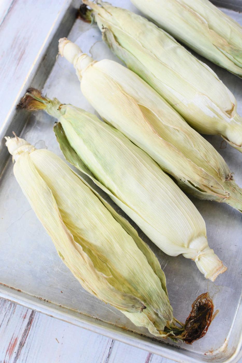Five unpeeled ears of corn on the cob rest side by side on a metal baking tray, ready for oven cooking, arranged neatly on a wooden surface.