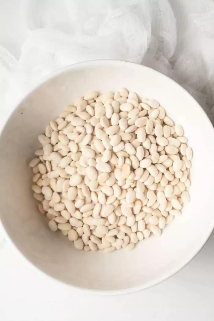 A white bowl filled with dried Great Northern beans is placed on a light-colored surface.