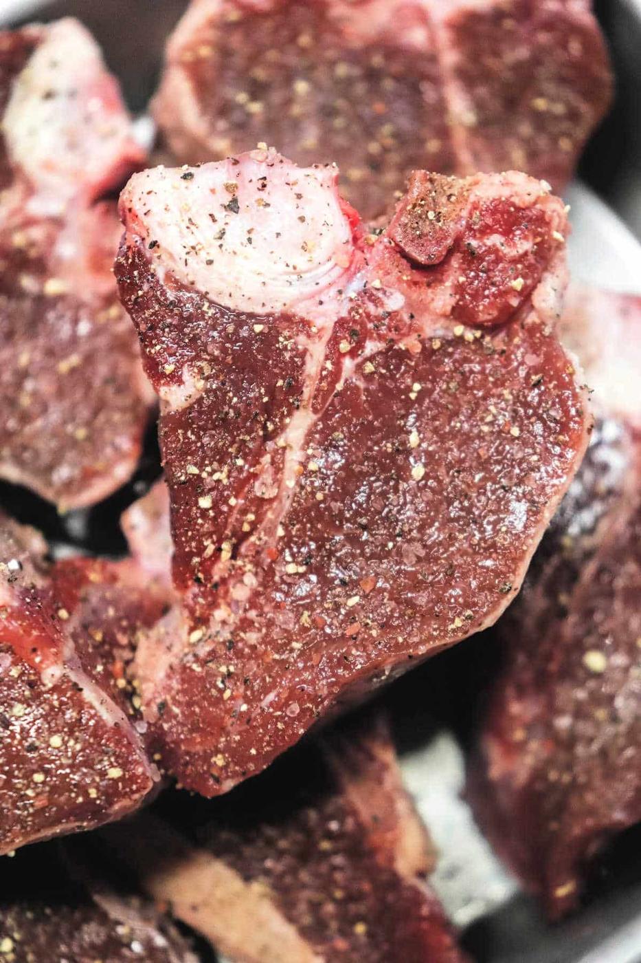 Close-up of raw, seasoned lamb loin chops piled together. The meat is seasoned with visible black pepper and salt, similar to the methods used in How to Cook Lamb Loin Chops. The texture of the steaks is marbled with some fat and dark red lean meat.