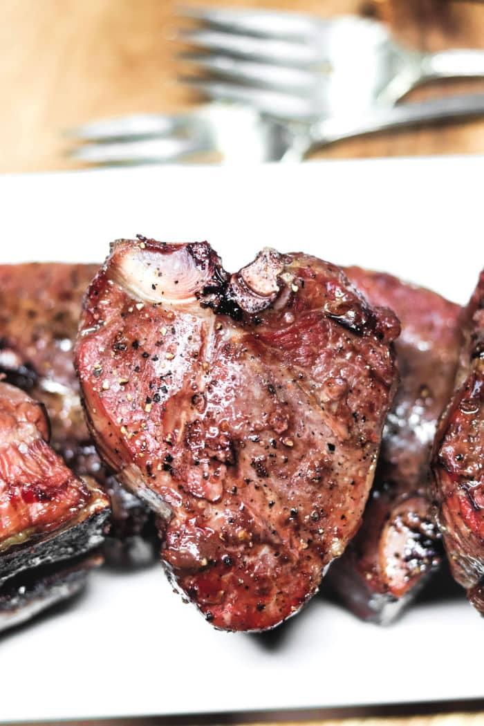 Close-up of seasoned, grilled lamb loin chops on a white plate with blurred forks in the background, offering a visual delight akin to mastering how to cook lamb loin chops.