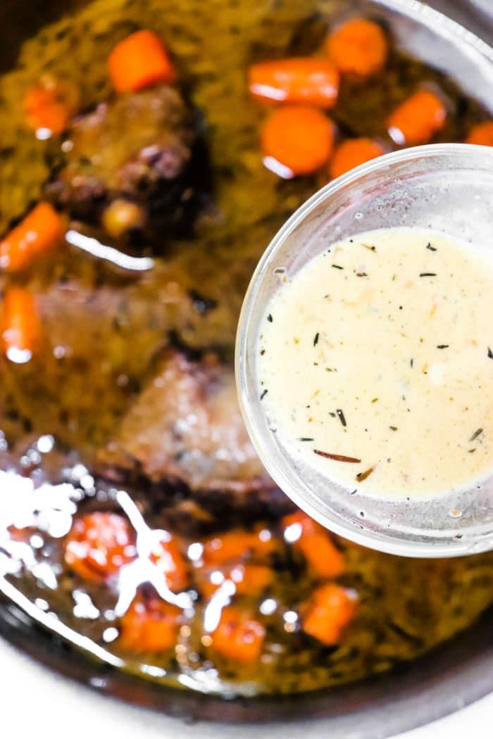 A dish with tender, cooked oxtail pieces and sliced carrots in a dark sauce is shown in a pan, reminiscent of how to cook oxtails on the stove. Above the pan, a small glass bowl holds a light creamy sauce speckled with visible herbs.
