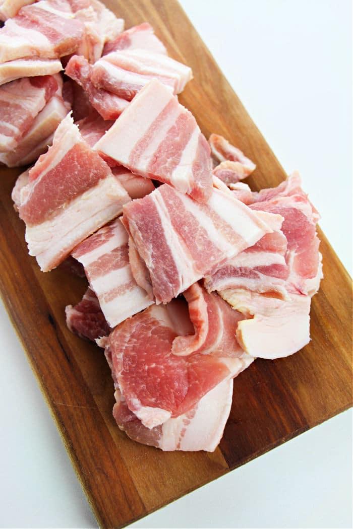 Raw bacon pieces, ideal for learning how to cook pork belly in the oven, are stacked on a wooden cutting board against a white background.