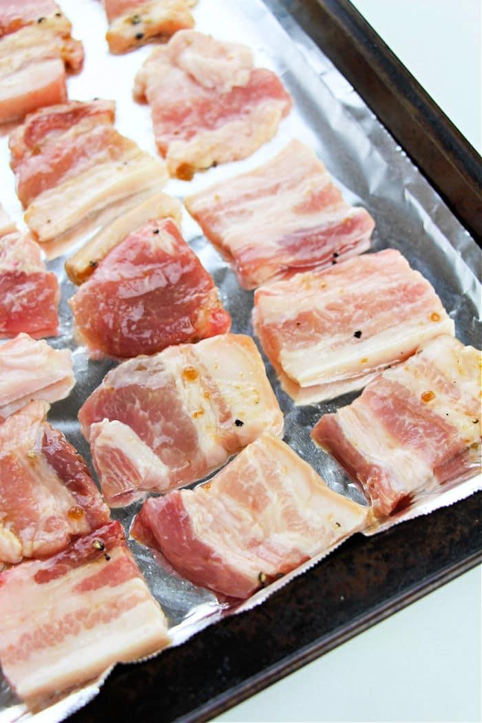 Raw slices of pork belly arranged on a foil-lined baking sheet, ready for the next step in learning how to cook pork belly in the oven.