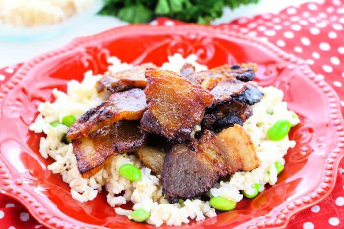 Crispy bacon pieces—perfect for those learning how to cook pork belly in the oven—are served on a bed of rice with green edamame on a red plate.
