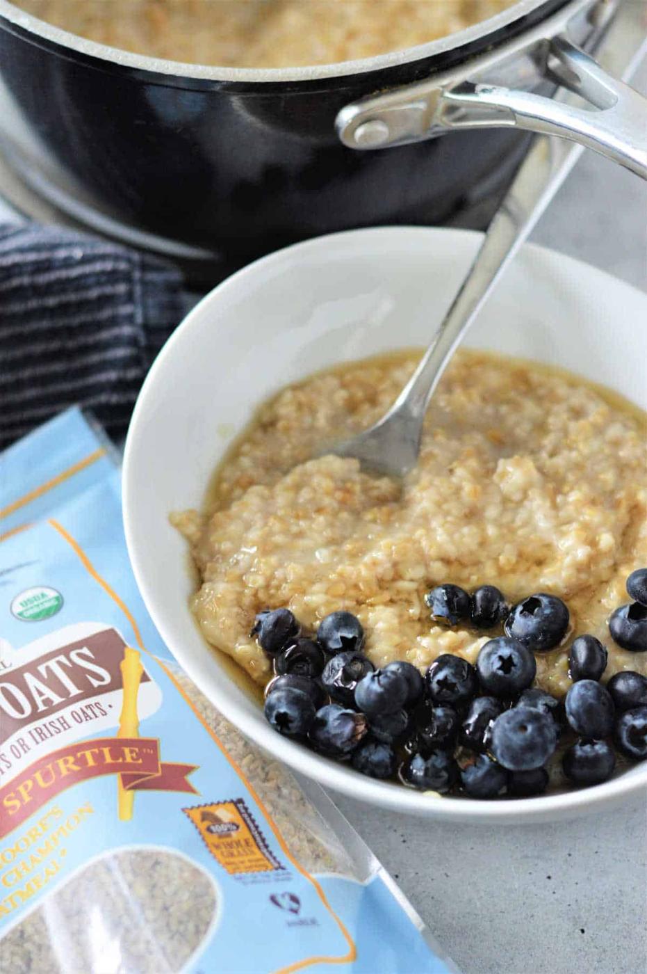 Bowl of steel cut oatmeal with blueberries, next to a package of steel-cut oats and a pot in the background, illustrating how to cook steel-cut oats.