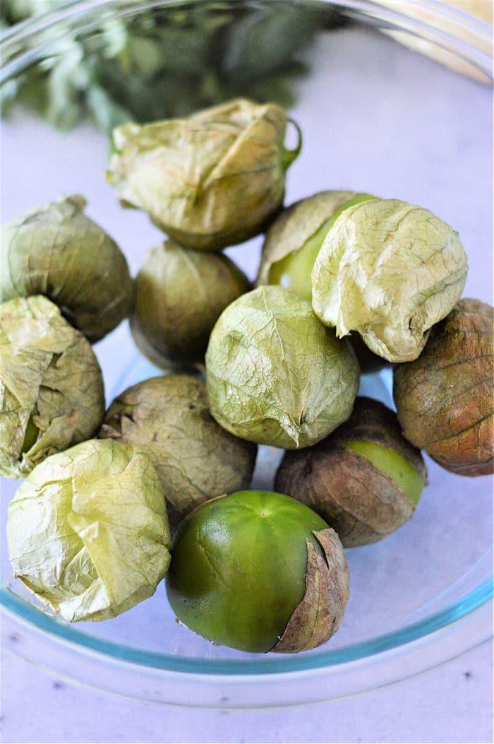 Tomatillos in husks rest inside a glass bowl, with leafy greens subtly framing them in the background, inviting you to explore how to cook tomatillos