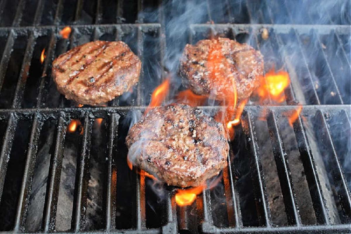 Three beef patties grilling over open flames and smoke on a barbecue grill—discover how to grill frozen burgers to perfection with this mouthwatering scene.