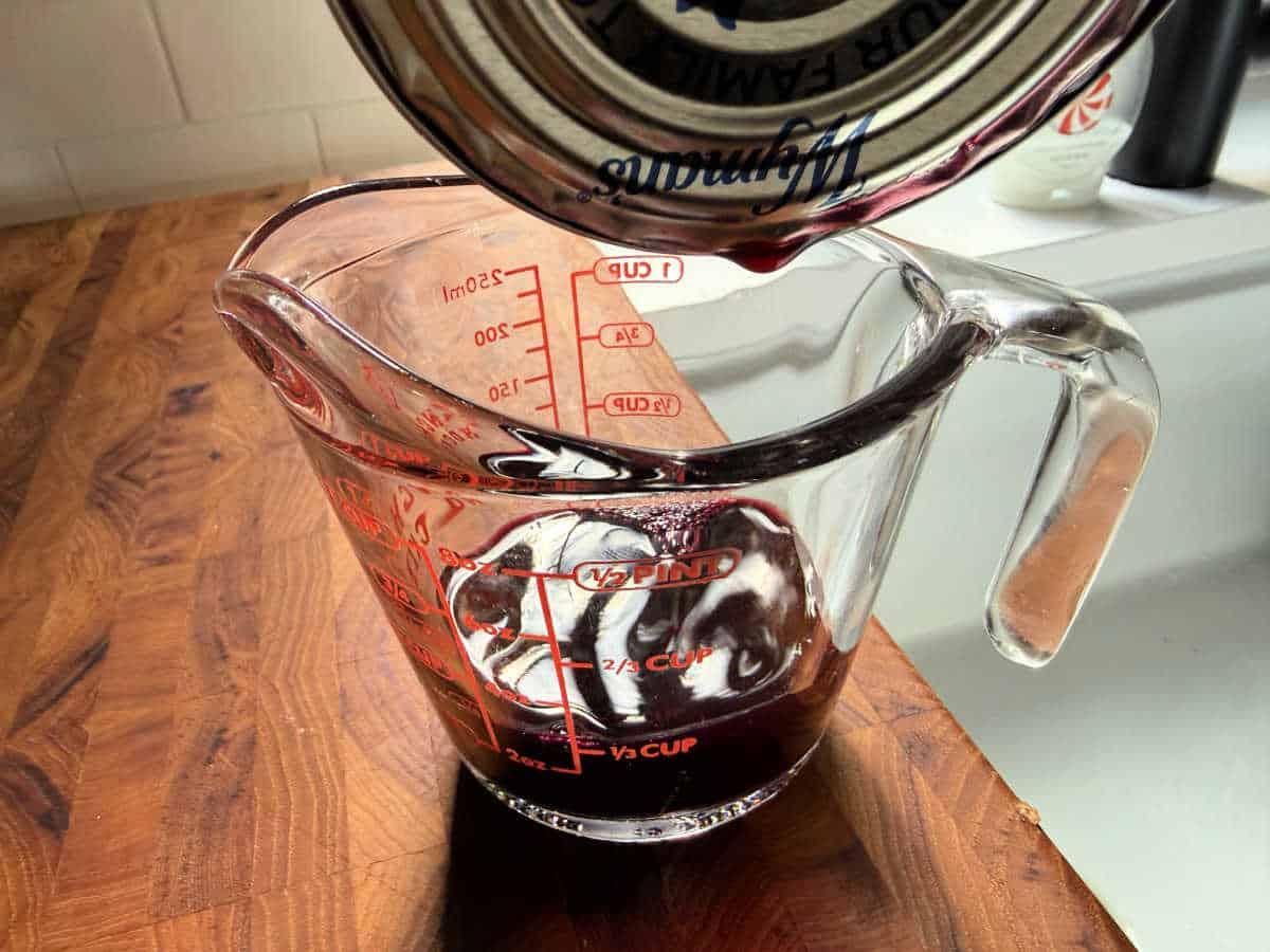 Red liquid being poured from a can into a glass measuring cup on a wooden countertop—perfect for learning how to make blueberry muffin mix better with flavorful additions.