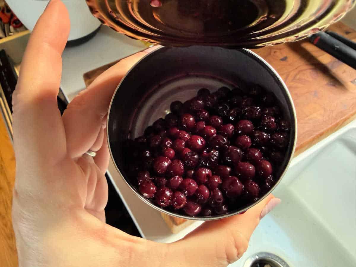 A hand holds an open can filled with dark purple berries over a kitchen sink and cutting board—perfect for learning how to make blueberry muffin mix better.