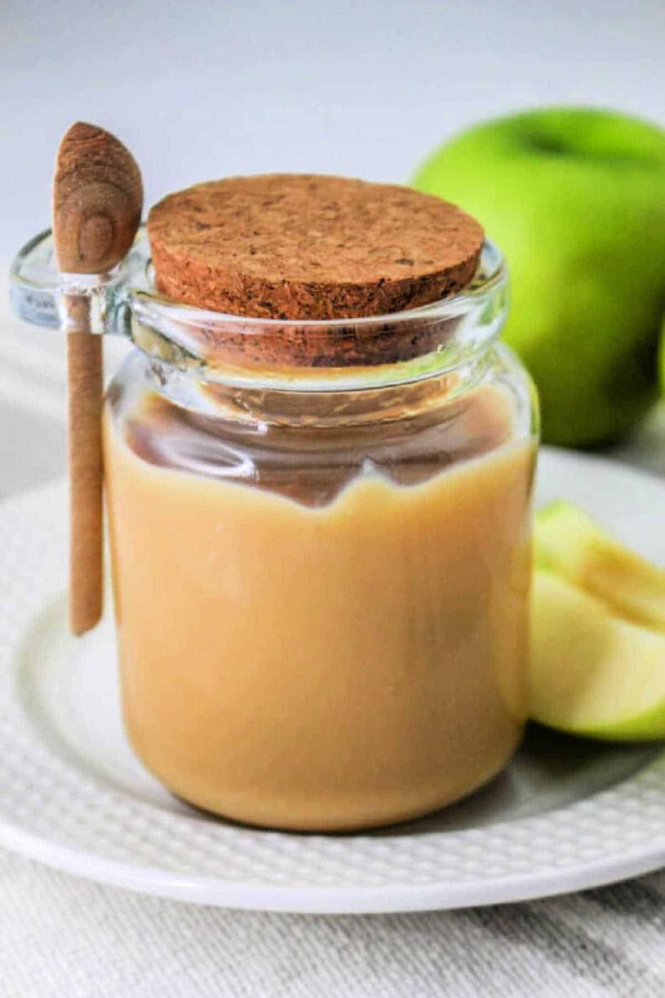 Glass jar of caramel sauce made with condensed milk, cork lid, and a small wooden spoon, next to apple slices on a white plate—perfect for those learning how to make caramel with condensed milk at home.