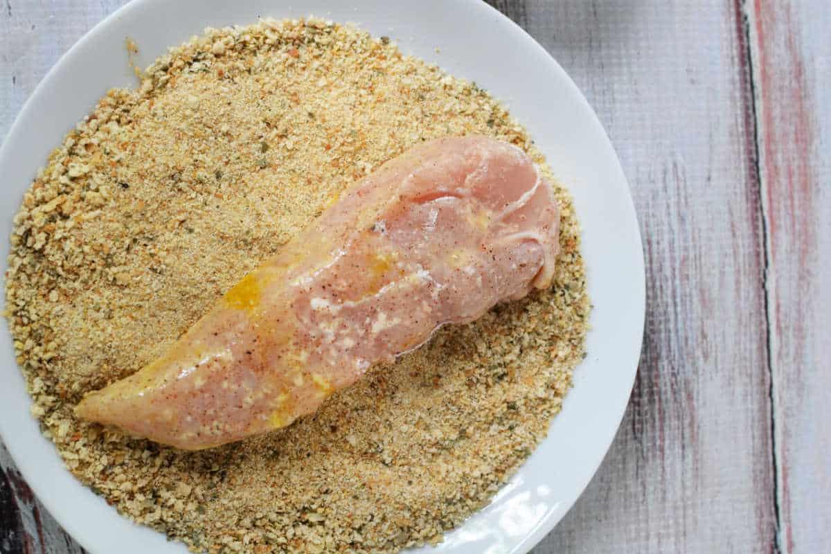 Learn how to make chicken tenders by breading raw chicken breast on a white plate, achieving that perfect crispy texture.