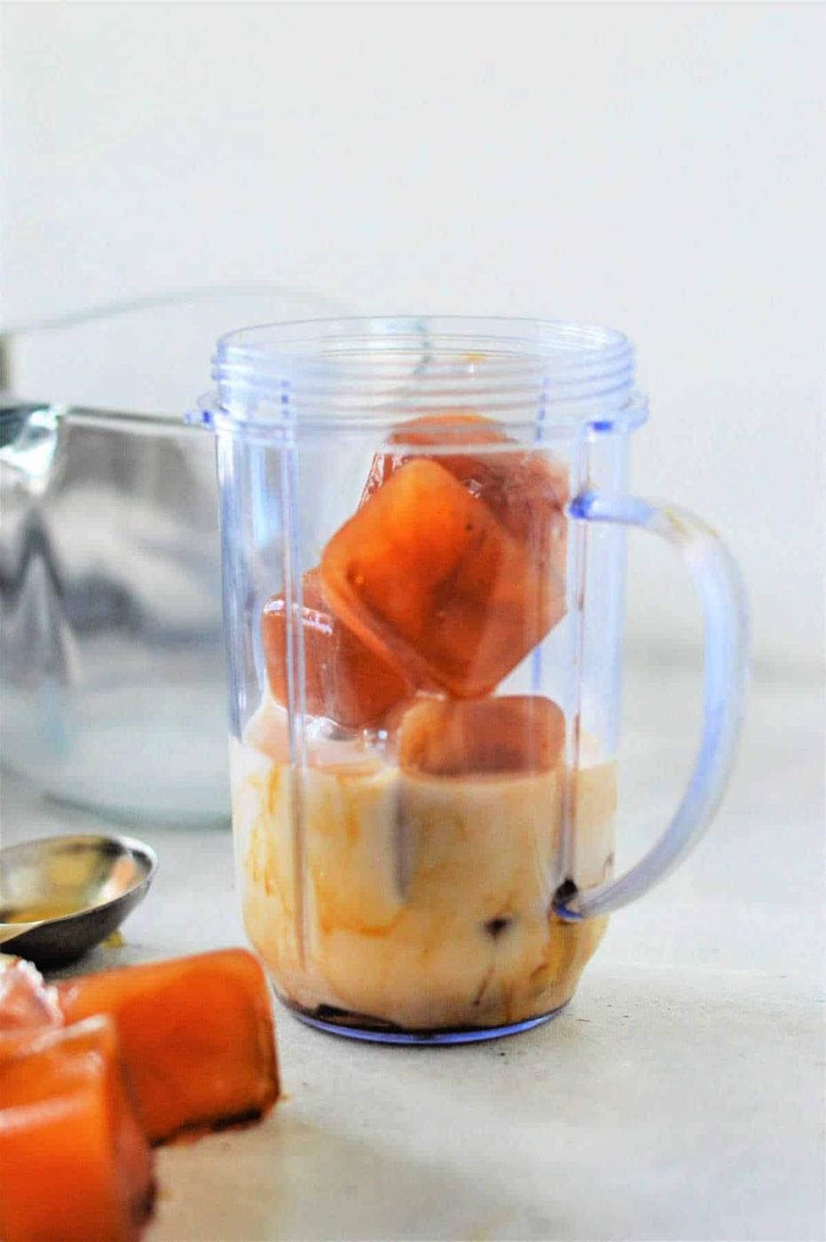 A plastic blender cup is filled with milk and several coffee ice cubes, illustrating how to make cold foam for coffee. A metal spoon and another coffee ice cube rest on the surface beside the blender cup, with a larger mixing container visible in the background.