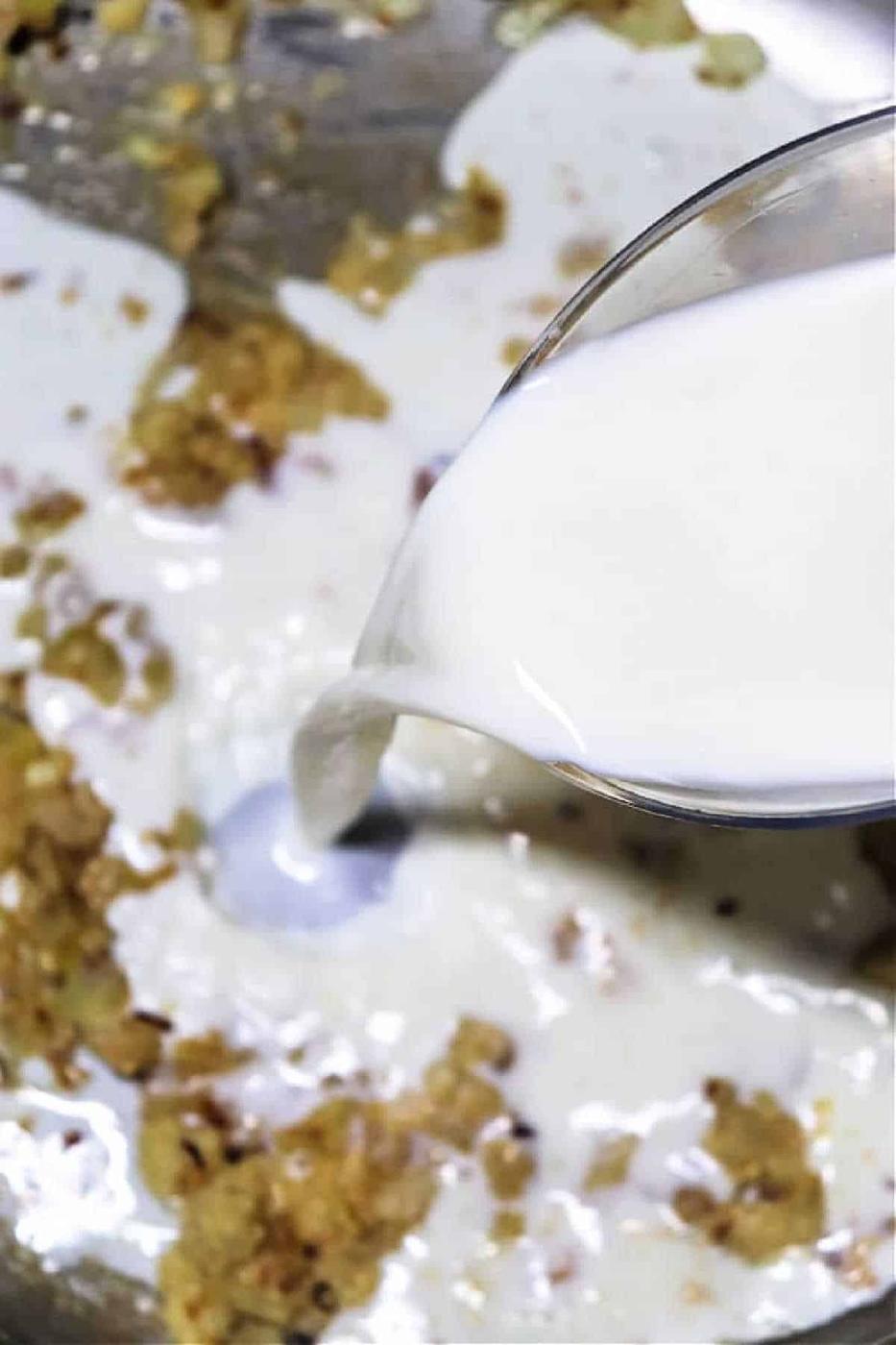 Milk is poured from a glass jug into a pan with sautéed onions and spices, showing an essential step in how to make gravy from chicken drippings for a rich, creamy finish.