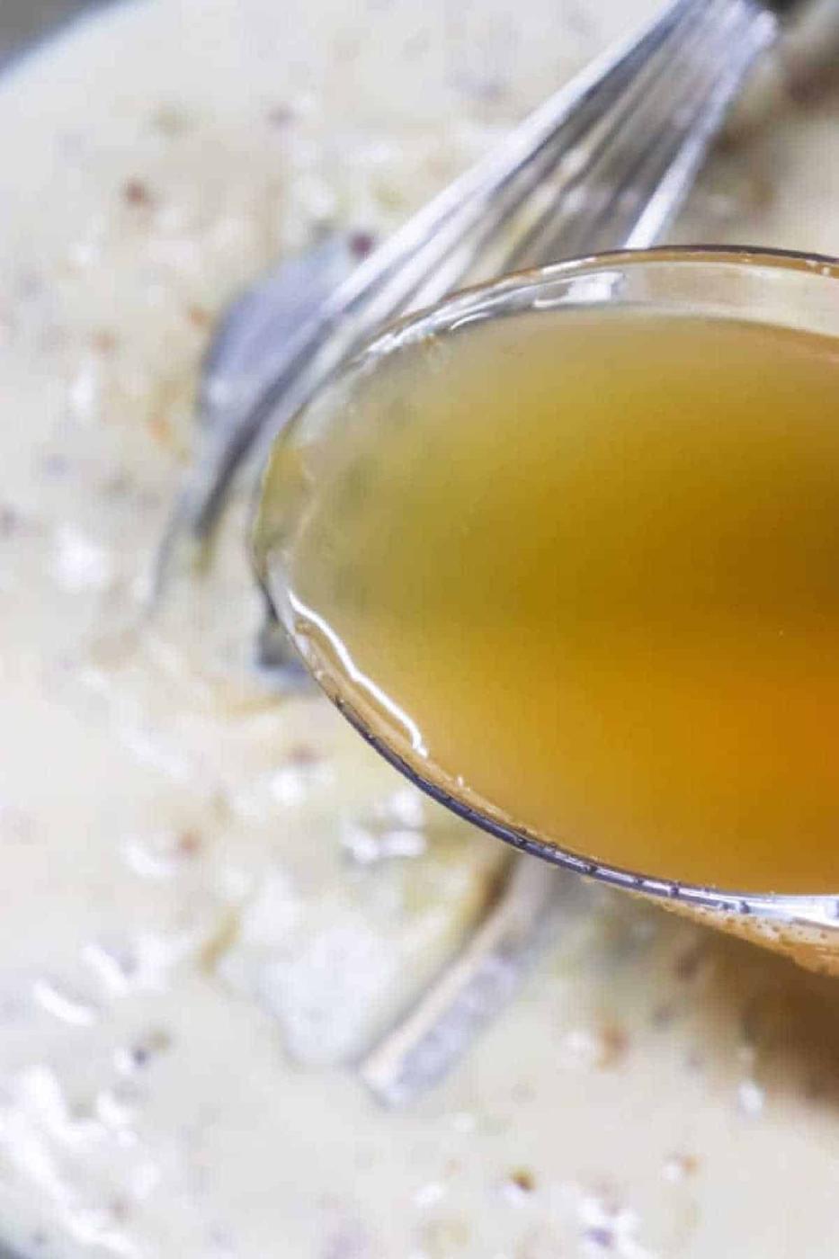 A spoon with clear broth held above creamy soup with a whisk in the background, perfect for illustrating how to make gravy from chicken drippings.