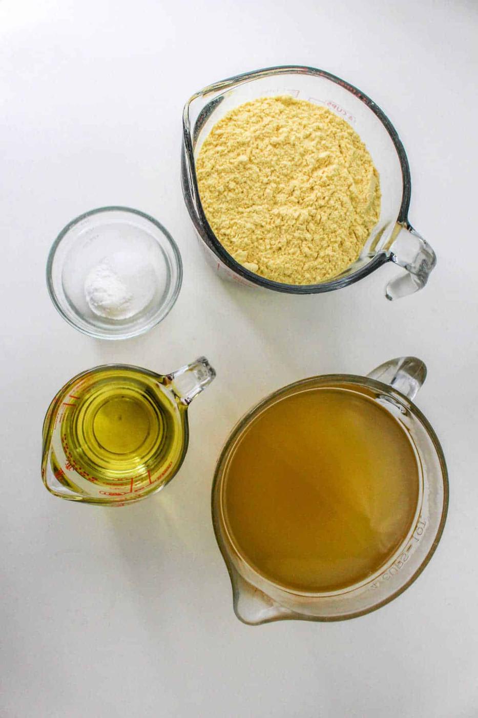 Four measuring cups filled with cornmeal, oil, broth, and salt are arranged on a white surface, showcasing the essential ingredients for how to make tamales masa.