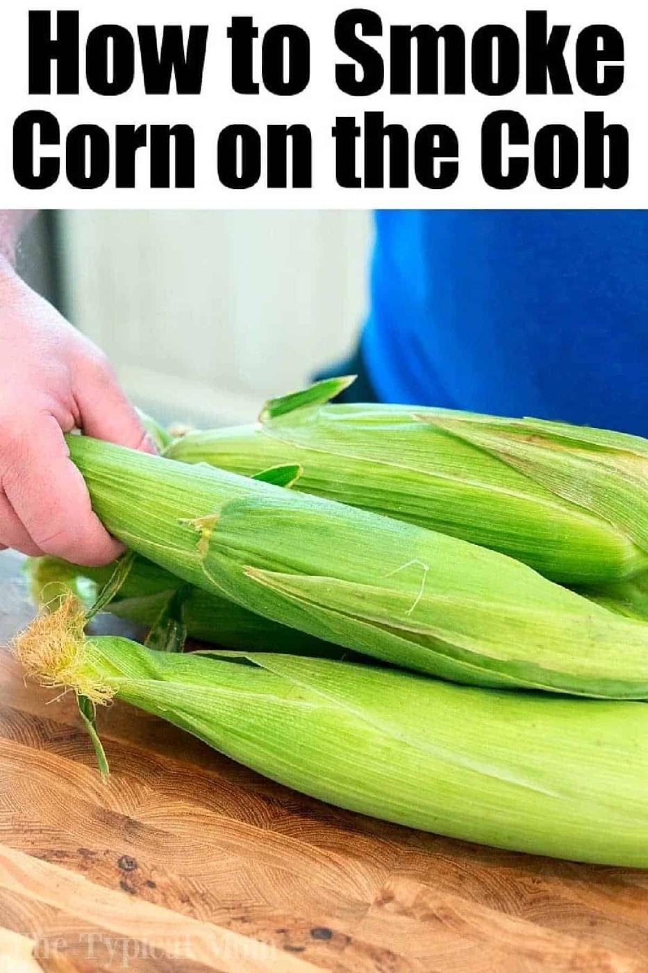 Hand holding fresh ears of corn in husks on a wooden surface, with text: Discover how to smoke corn on the cob for a delicious, smoky flavor.