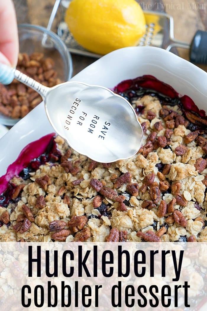 A spoon rests beside a generous serving of huckleberry cobbler in a white dish, with the words SAVE ROOM FOR SECONDS inviting you to enjoy every delicious bite.