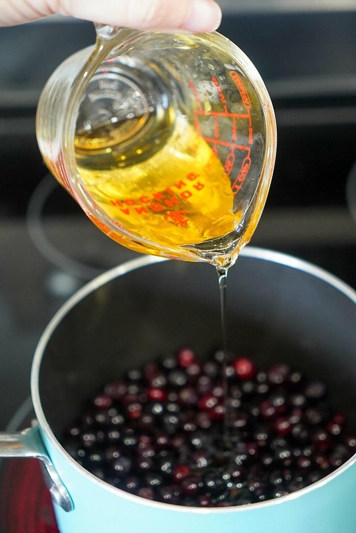 Pouring honey into a saucepan filled with huckleberries on a stovetop.