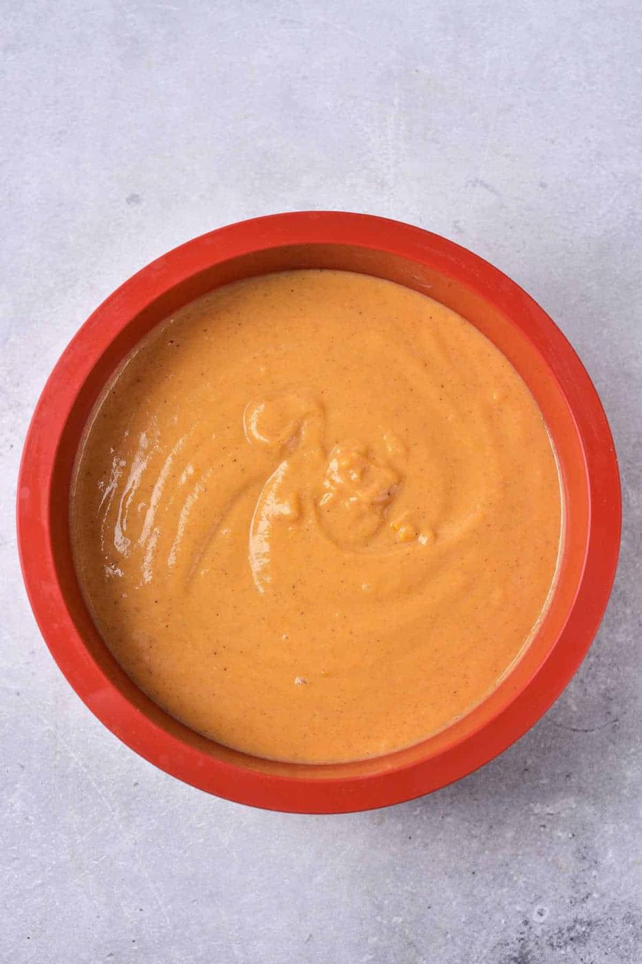 A red bowl filled with creamy, light brown Impossible Pumpkin Pie sauce sits on a gray, textured surface.