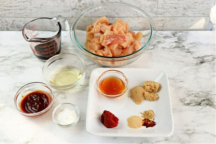 Various ingredients in bowls and a plate, including diced raw asian chicken, sauces, oil, and assorted spices on a marble surface.