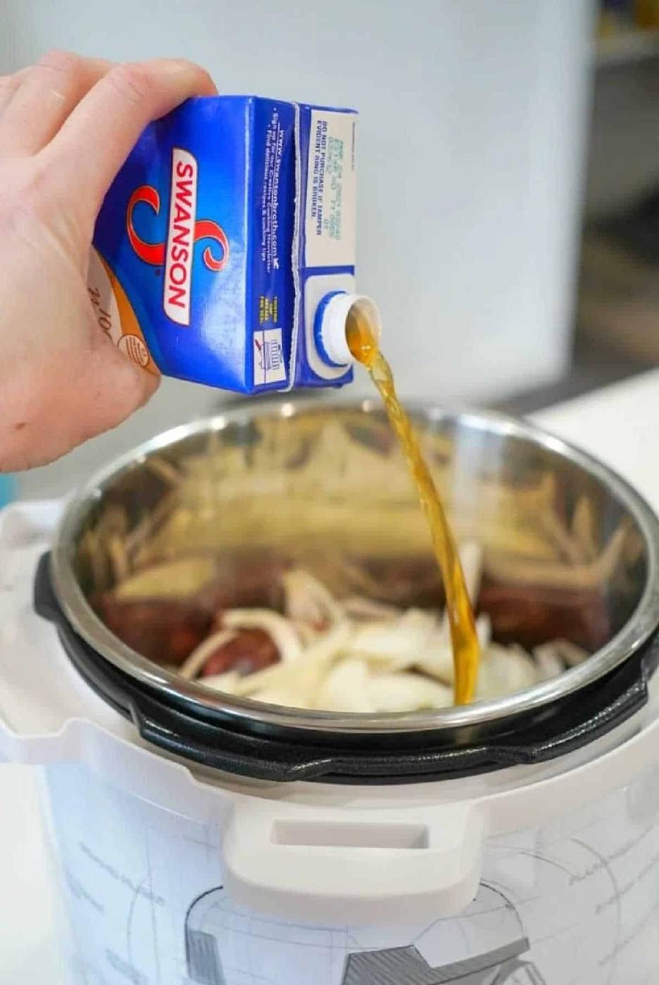 A hand pours Swanson broth from a carton into a pressure cooker filled with sliced onions, prepping for delicious instant pot BBQ beef.