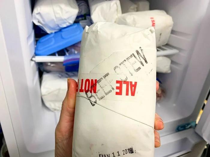 A hand holds a white paper-wrapped package labeled BEEF STEW MEAT, perfect for an instant pot recipe, with a date stamp. Other similarly wrapped packages and a blue ice pack are visible in the freezer in the background.