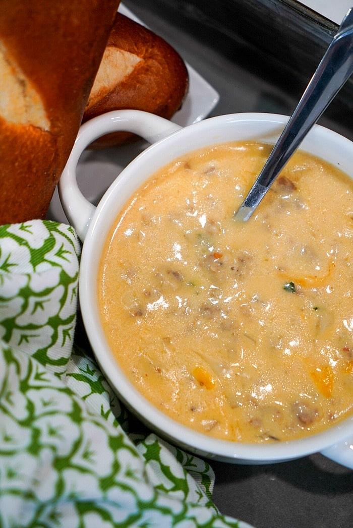A bowl of creamy beer cheese soup with visible chunks, a spoon inside, bread, and a green patterned cloth nearby evokes the comforting scent of an Instant Pot creation.