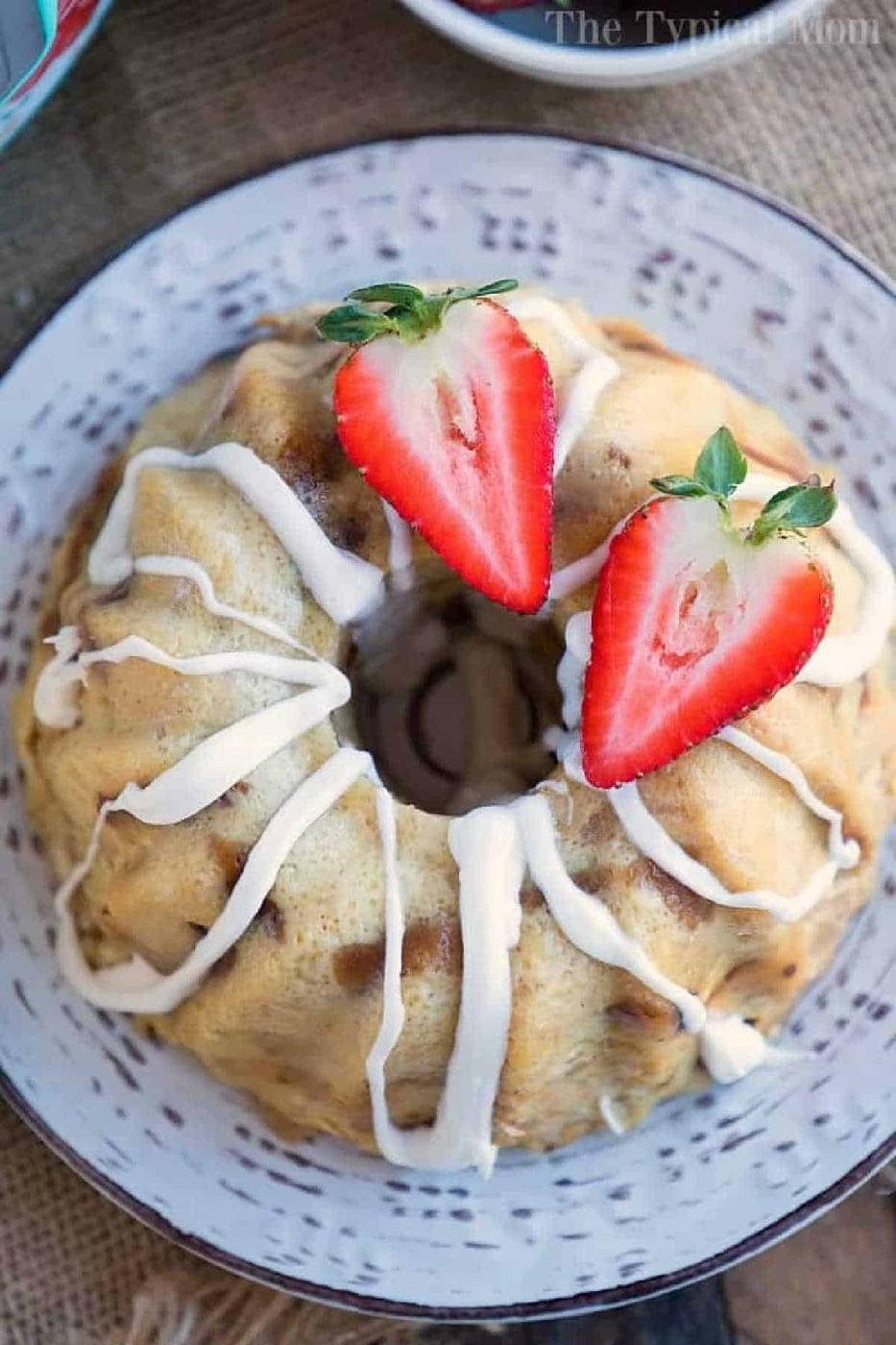 A bread pudding bundt cake with white icing, topped with halved strawberries, sits gracefully on a decorative plate, reminiscent of the cozy warmth that instant pot bread pudding brings to a home.