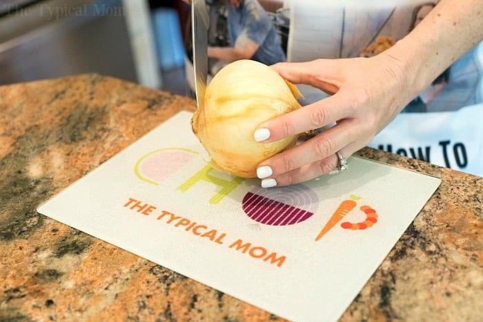 A person with white nail polish is slicing an onion on a cutting board marked with "CHOP Zopago," preparing ingredients for an instant pot cheeseburger soup. The cutting board rests elegantly on a granite countertop.