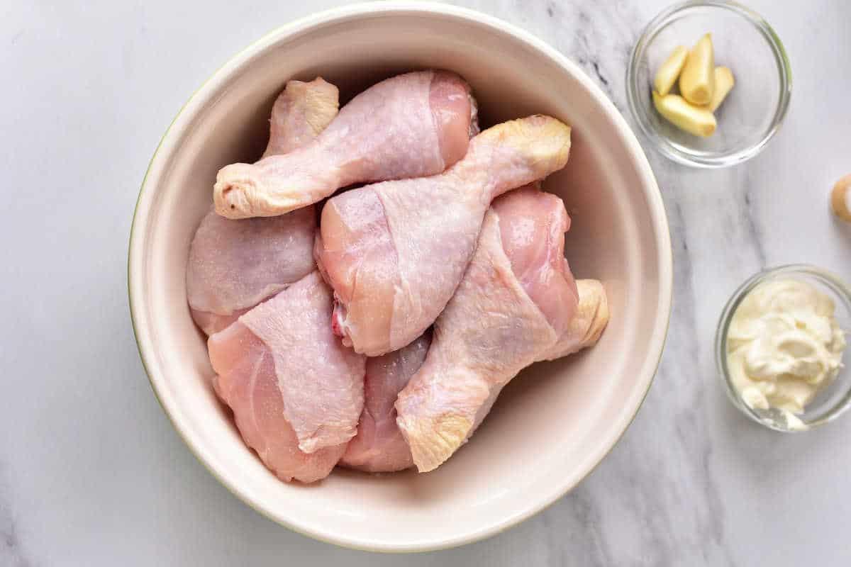 Raw chicken drumsticks in a bowl, with garlic cloves and mayonnaise in small bowls beside it on a white surface—perfect for making flavorful instant pot chicken legs.