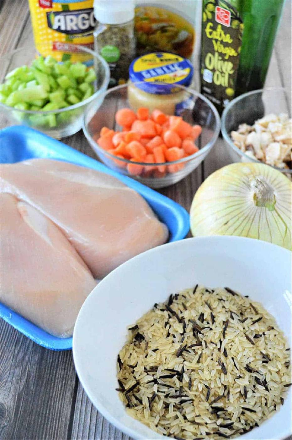 Raw chicken breasts, wild rice, chopped carrots, celery, onion, and various seasonings are ready on a wooden table—perfect ingredients for a cozy instant pot chicken rice soup.