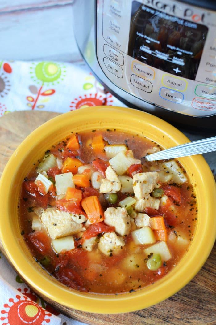 A yellow bowl brimming with Instant Pot chicken vegetable soup features tender chicken, diced potatoes, carrots, peas, and tomatoes. A spoon rests in the bowl while an Instant Pot sits in the background on a floral patterned cloth.