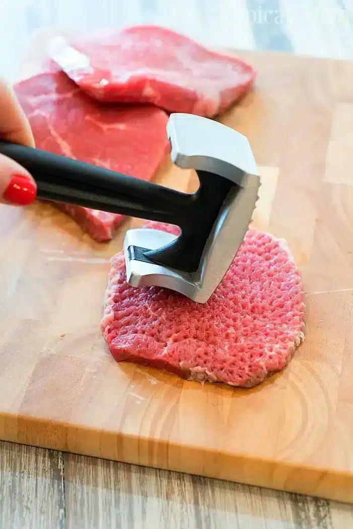 A person uses a meat tenderizer to pound a slice of raw beef on a wooden cutting board, preparing it for a Ninja Foodi cube steak recipe. Two more slices of raw beef are visible in the background on the cutting board.