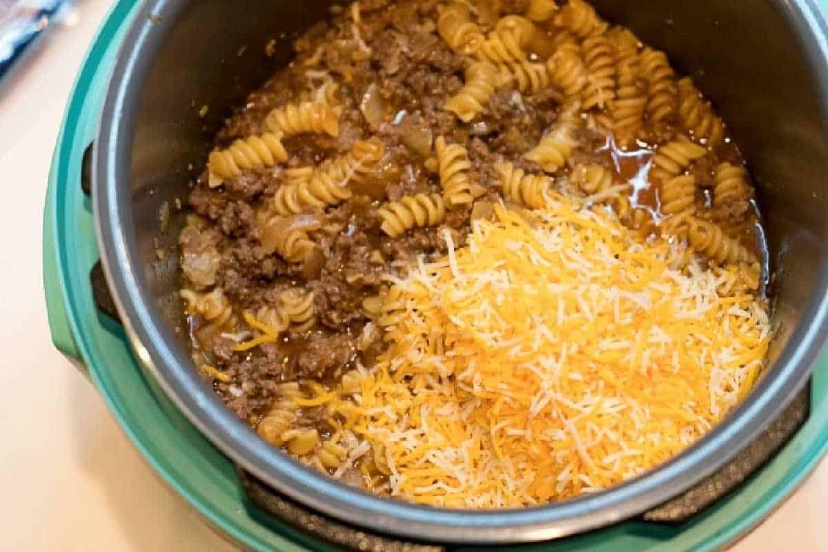 A pot with cooked rotini pasta, ground beef, and a pile of shredded cheese on top—perfect for transforming into an Instant Pot enchilada casserole.
