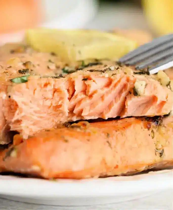 A close-up of a piece of cooked salmon on a plate with herbs and a lemon slice, prepared perfectly from frozen in an Instant Pot. A fork is placed into the flaky, pink meat, highlighting its texture.