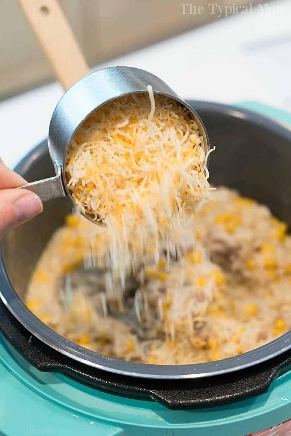 A hand pours shredded cheese from a measuring cup into a pot of rice and corn, just like preparing an instant pot hamburger casserole.