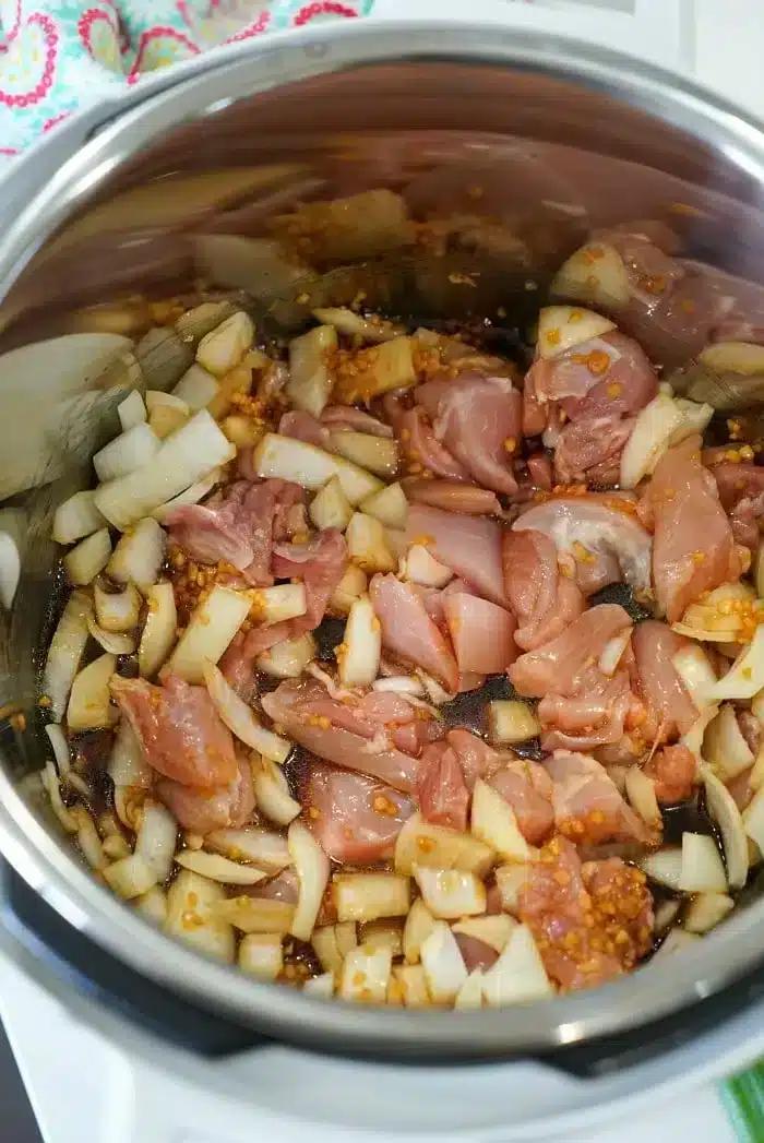 A metal pot contains uncooked diced chicken, chopped onions, and minced garlic. The ingredients are in the process of being sautéed, with some onions already starting to turn translucent, creating the base for a delicious Instant Pot Korean chicken dish.