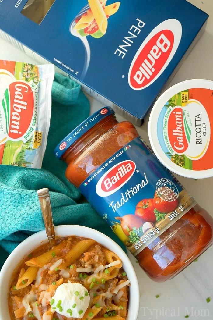 A colorful flat-lay showcasing ingredients for a pasta dish: a box of Barilla penne, a jar of Barilla Traditional pasta sauce, a container of Galbani ricotta cheese, and a package of Galbani mozzarella cheese. A prepared bowl of instant pot lasagna soup is also included.
