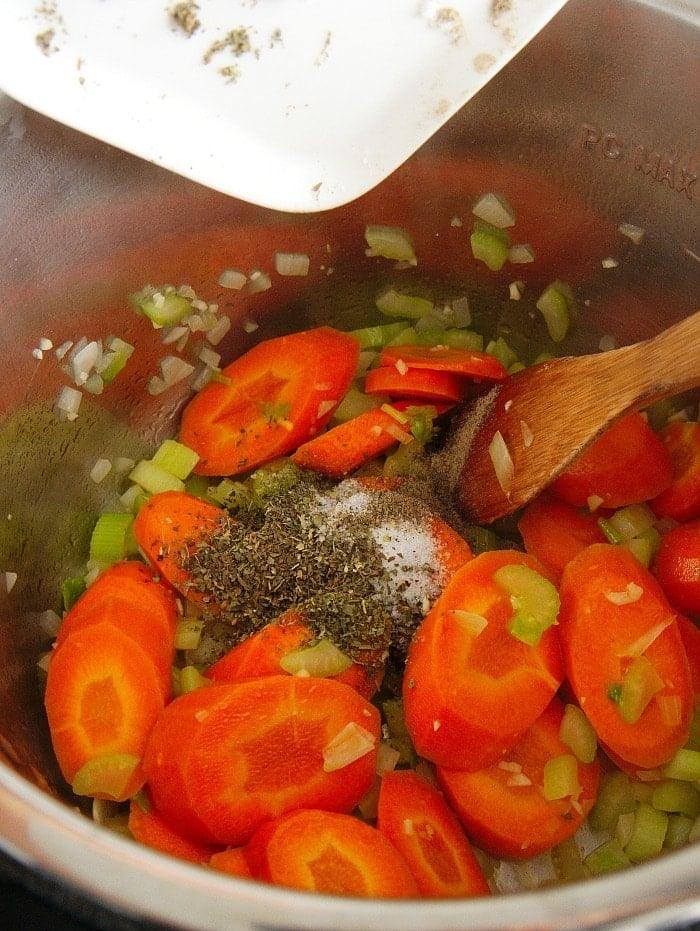 Sliced carrots, celery, onions, and seasonings being stirred with a wooden spoon in a metal pot for instant pot meatball soup.