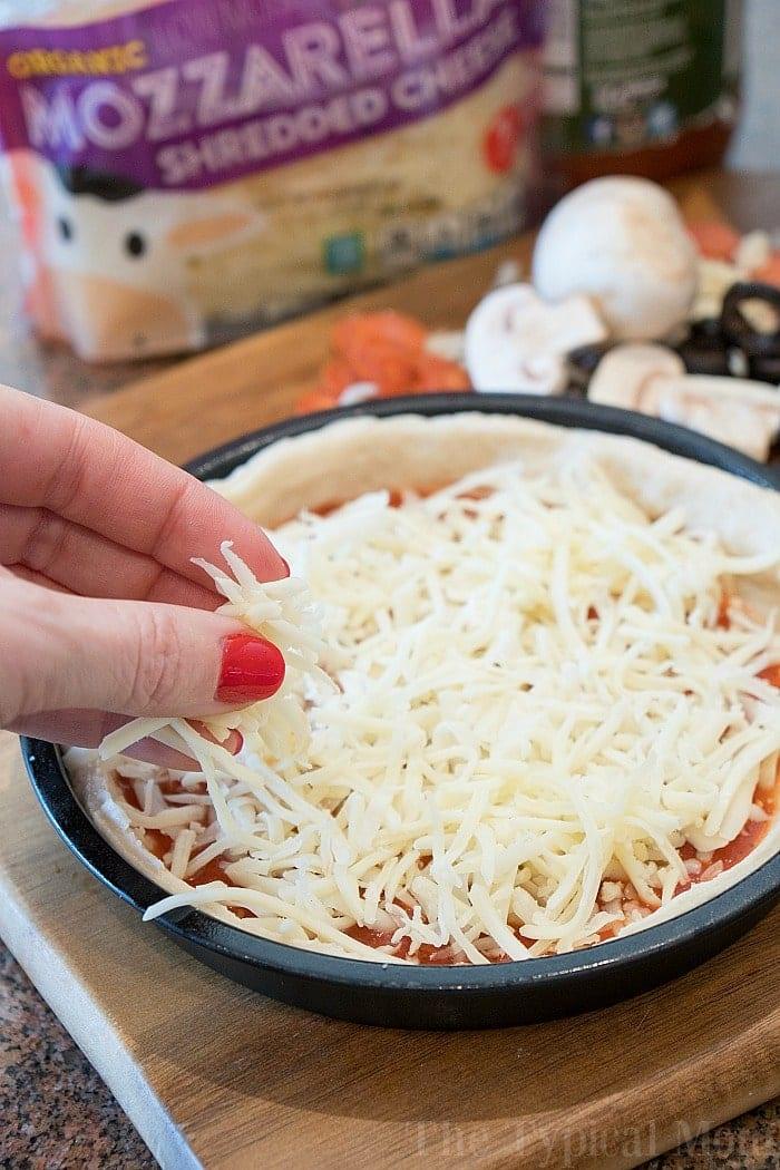 A hand sprinkles shredded mozzarella cheese onto a pizza crust with sauce, ready to be transformed into a delicious instant pot pizza. Mushrooms and olives nearby await their turn as tasty toppings.