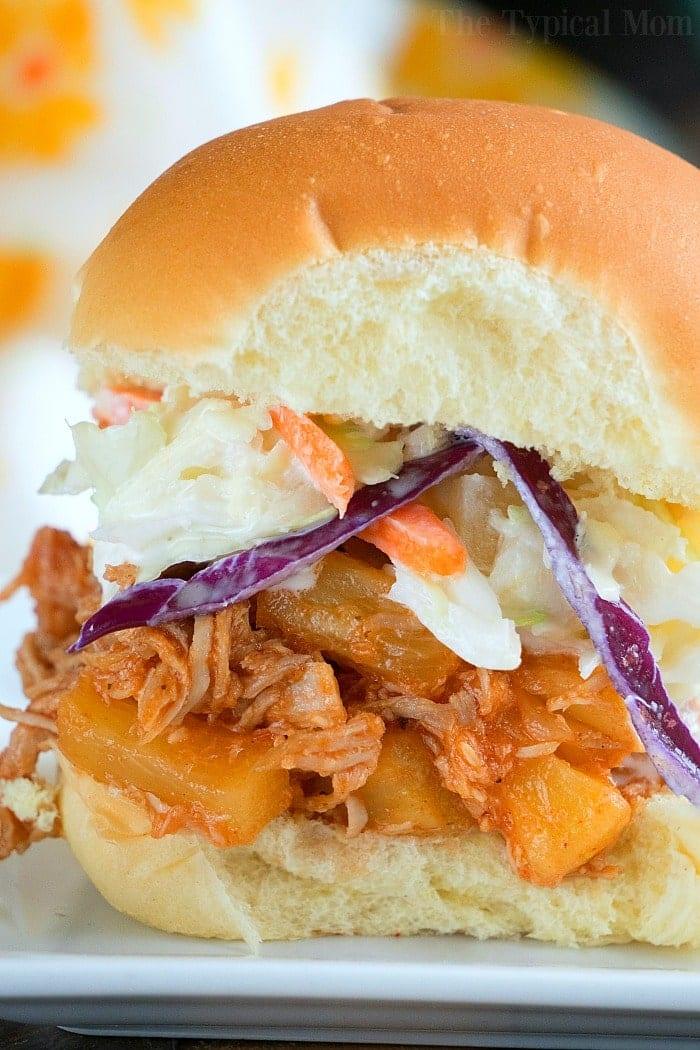 A close-up of an Instant Pot Hawaiian pulled pork sandwich, featuring tender shredded meat, pineapple pieces, and coleslaw inside a soft bun. The layers of colorful ingredients are visible, with vibrant purple cabbage and carrot strands enhancing the coleslaw.