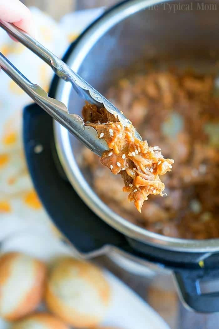 Tongs hold a portion of teriyaki pulled pork over an open Instant Pot. The succulent meat is mixed with sauce and sprinkled with sesame seeds. A blurred background reveals bread rolls and part of a printed cloth, adding a cozy touch to this culinary scene.